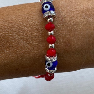 May include: Red beaded bracelet with two blue evil eye charms. The charms are surrounded by clear rhinestones.