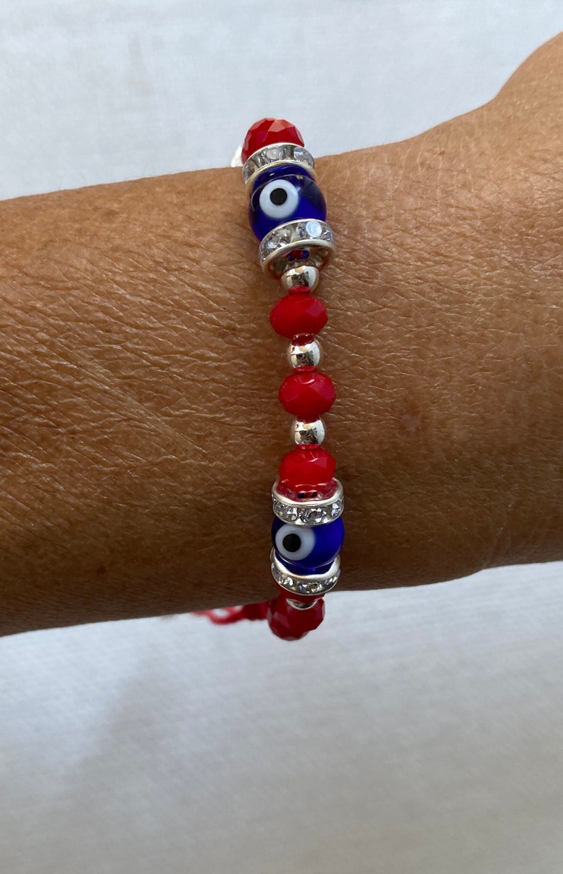 May include: Red beaded bracelet with two blue evil eye charms. The charms are surrounded by clear rhinestones.