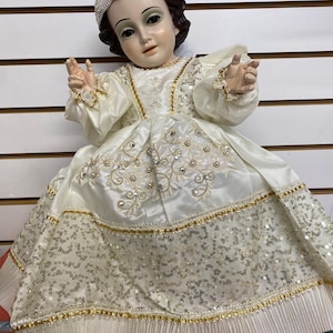 May include: A doll of a baby Jesus figure wearing a white satin dress with gold trim and embellishments. The dress is adorned with sequins and pearls. The doll has dark hair and is wearing a crown.