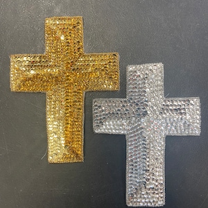 May include: Two sequined crosses, one gold and one silver, are displayed against a dark background. The crosses are made of small, reflective sequins, giving them a shimmering appearance. The gold cross is on the left, and the silver cross is on the right.