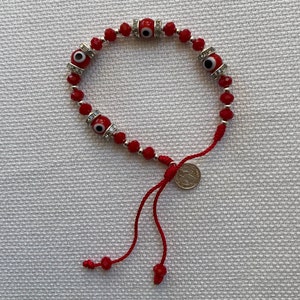 May include: Red beaded bracelet with four silver evil eye charms and a silver coin charm. The bracelet has a red string tie closure.