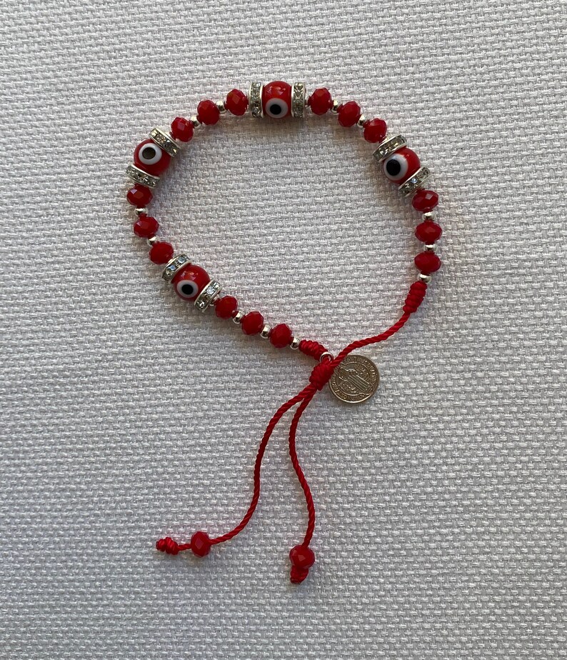 May include: Red beaded bracelet with four silver evil eye charms and a silver coin charm. The bracelet has a red string tie closure.