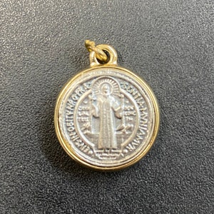 May include: A gold-toned pendant with a silver-toned Saint Anthony medal. The medal features a depiction of Saint Anthony holding a book and a lily. The inscription around the medal reads "S. Antonius de Padua" and "Ora pro nobis".