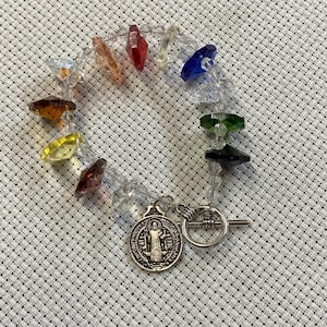May include: A beaded bracelet featuring a spectrum of colorful, faceted crystals. The bracelet includes red, orange, yellow, green, and blue crystals, strung on a clear cord. A silver-toned charm with a religious figure and a toggle clasp complete the design.
