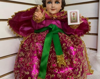 May include: A small, seated figurine of the baby Jesus wearing a pink and gold dress with a green sash. The figurine has a halo and is holding a small card with a picture of the baby Jesus.