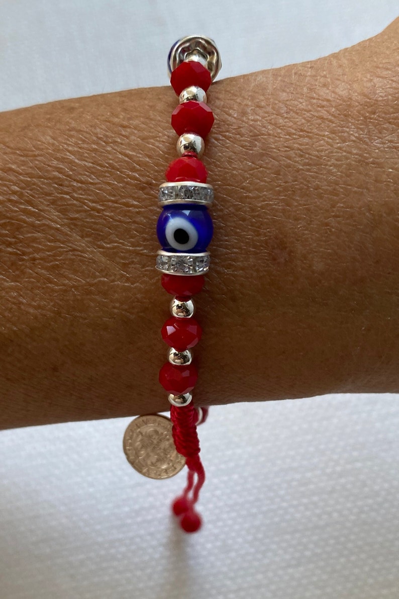May include: Red string bracelet with a blue evil eye charm and silver beads. The bracelet has a silver coin charm at the end.