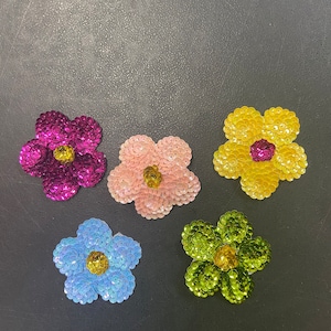 May include: Five fabric flower patches with sequin embellishments. The flowers are in shades of pink, yellow, blue, and green. The centers of the flowers are yellow.
