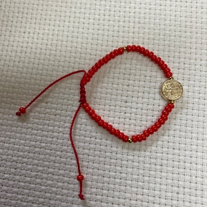 May include: A red beaded bracelet with a gold-colored medallion. The bracelet is made of small, round red beads and features a gold-colored medallion with embossed details. The bracelet has a red string closure.