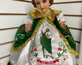 Puede incluir: Una pequeña muñeca blanca y verde vestida con un vestido de satén blanco con ribetes de terciopelo verde y una capa de terciopelo verde. La muñeca tiene una peluca marrón y sostiene un bastón. La muñeca está bordada con las palabras "San Judas".