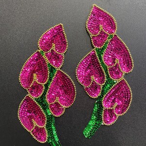 May include: Two pink and green sequined flower appliques. Each applique features a cluster of pink heart-shaped sequins with green sequins outlining the petals and stems.