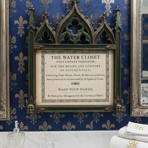 Pode incluir: Uma placa emoldurada com a inscrição "THE WATER CLOSET" com detalhes ornamentais verdes e prateados, montada em uma parede azul com um padrão de flor-de-lis. Abaixo, toalhas brancas com iniciais bordadas estão empilhadas. O texto está em escrita inglesa antiga.