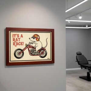 Può includere: Una stampa incorniciata con un topo dei cartoni animati su una moto rossa. Il topo indossa un casco e occhiali da sole. Il testo "IT'S A RAT RACE!" è sopra l'immagine. La cornice è marrone scuro.