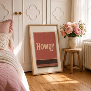 May include: A framed wall art print featuring a vintage-style red matchbox graphic with the word HOWDY printed in cream-coloured lettering. The design includes a dark brown strike strip at the bottom of the matchbox illustration.