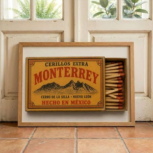 May include: A framed print of a vintage matchbox labeled Cerillos Extra Monterrey, featuring a mountain illustration, red text, and a partially open drawer revealing wooden matches with orange tips, set in a light wood frame.