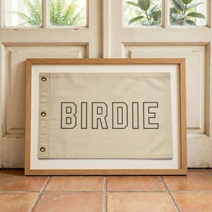 Puede incluir: Una bandera rectangular de lona beige con la palabra BIRDIE en letras negras de bloque. La bandera tiene tres ojales metálicos de color latón en el lado izquierdo y se muestra dentro de un marco de madera clara con un borde de paspartú blanco.