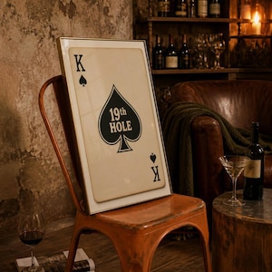 Puede incluir: Una pieza de arte mural rectangular enmarcada que presenta un diseño de carta de juego de estilo vintage. La carta es un Rey de Picas con un fondo color crema, un símbolo de pica negro y el texto 19th HOLE impreso en el centro del símbolo.