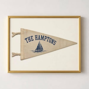 Pode incluir: Uma flâmula triangular de feltro bege com o texto em azul escuro THE HAMPTONS e um gráfico de um veleiro. A flâmula tem dois pequenos laços no lado esquerdo e é exibida dentro de uma moldura retangular fina de cor dourada.