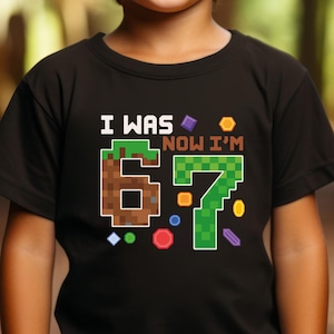 May include: Black t-shirt with the text "I WAS 6 NOW I'M 7" in pixelated font. The number 6 is brown with a green top, and the number 7 is green. Colorful pixelated shapes surround the numbers, suitable for a child's birthday.