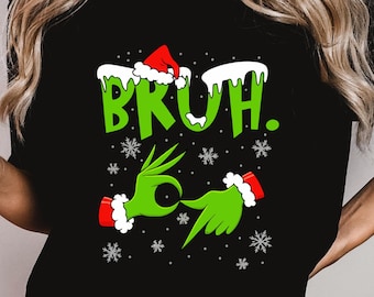 May include: Black t-shirt with a green graphic of a hand gesture and the word "BRUH" in green. The design includes snowflakes and a red Santa hat. The hand gesture is a green Grinch-like hand.