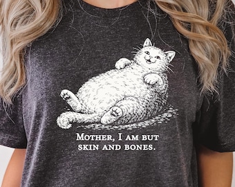 May include: Dark gray t-shirt featuring a white illustration of a cat lying on its back with the text "MOTHER, I AM BUT SKIN AND BONES." The cat illustration is detailed and whimsical, with a smiling expression.