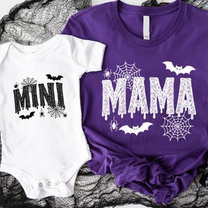 May include: A white baby onesie with the word "MINI" in black dripping font, bats, spiders, and spiderwebs. Next to it is a purple t-shirt with the word "MAMA" in a similar font, bats, spiders, and spiderwebs. Halloween-themed apparel.