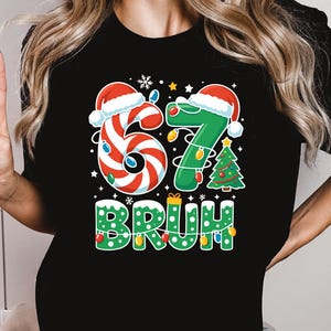 May include: Black t-shirt with a festive Christmas design. The design features the numbers "6" and "7" decorated with candy cane stripes and a Santa hat, along with a Christmas tree and the word "BRUH" in green lights.