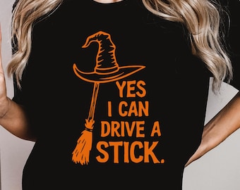 May include: Black t-shirt with an orange witch hat and broom graphic. The text reads, "YES I CAN DRIVE A STICK." The design is centered on the shirt, perfect for Halloween or witch-themed events.