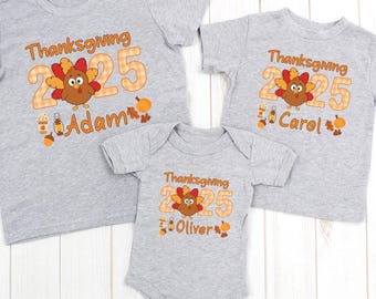 May include: Three gray t-shirts and a onesie with Thanksgiving-themed graphics. Each shirt features a turkey illustration, the year "2025", and a name. The graphics are in orange, brown, and yellow. The onesie has the same design.
