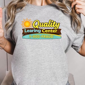 May include: A light gray t-shirt with the words "Quality Learing Center" in yellow and brown, with a sun graphic. Below the text is the phone number "1-800-FRAUD" in blue and white.