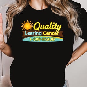 May include: Black t-shirt with a graphic that reads "Quality Learing Center 1-800-FRAUD". The design includes a yellow sun and text in yellow, brown, and blue. The shirt is a crew neck.