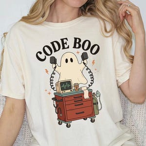 May include: A cream-colored t-shirt featuring a graphic of a ghost holding defibrillator paddles above a medical cart. The text CODE BOO is printed in black above the ghost. The cart includes a monitor, a coffee cup, and an oxygen tank.