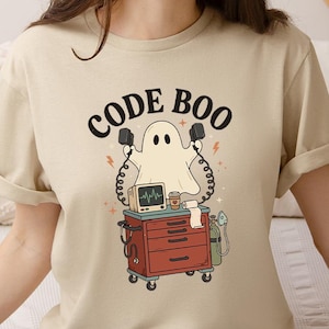 May include: A beige t-shirt featuring a graphic of a white ghost standing behind a red medical cart. The ghost holds two black defibrillator paddles. The text CODE BOO is printed above the ghost. The cart includes a monitor, coffee cup, and oxygen tank.