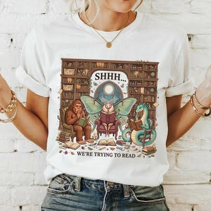 May include: A white short-sleeve t-shirt featuring a graphic of a Bigfoot, a moth with large green wings, and a green dragon sitting in chairs reading books in a library. The text reads SHHH... WE'RE TRYING TO READ.