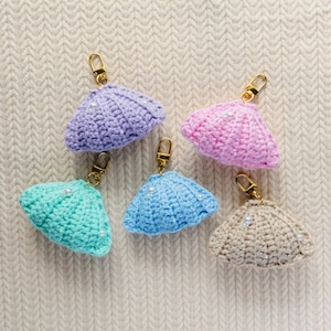 May include: Five handmade crochet seashell keychains in various colors: lavender, pink, teal, blue, and beige. Each shell is adorned with small white beads and a gold-colored clasp. The keychains are displayed on a textured, off-white surface.