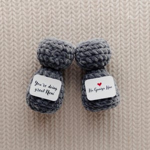 May include: Two gray, handmade, plush dolls with white tags. One tag reads "You're doing great Neni", the other reads "Hu Guaiya Hae" with a red heart. The dolls are on a beige, textured surface.