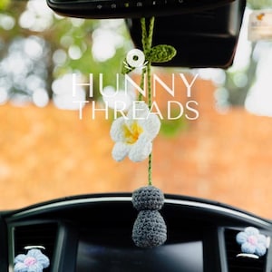 May include: A crochet car accessory hanging from a rearview mirror, featuring a white flower with a yellow centre, a green stem with leaves, and a dark grey textured base. The text HUNNY THREADS is overlaid in white across the centre of the image.