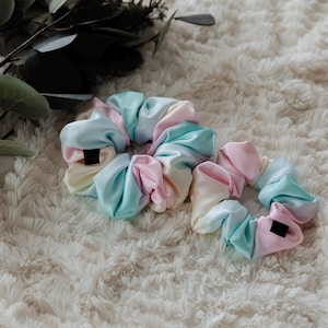 May include: Two pastel-colored scrunchies made of satin fabric. The scrunchies are in shades of pink, blue, and yellow. They are arranged on a white fluffy surface.
