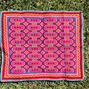 May include: A pink and blue embroidered throw pillow with a geometric pattern. The pillow has a red border and a blue and yellow inner border.