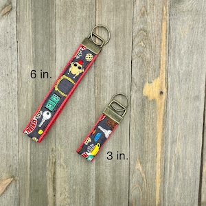 May include: Two keychains with a red and black fabric strap and a gold metal ring. The larger keychain is 6 inches long and the smaller keychain is 3 inches long.