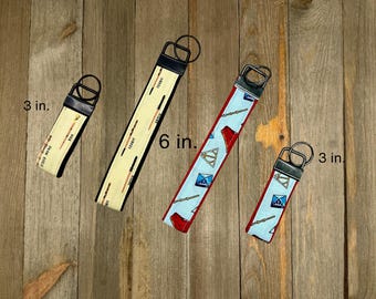 May include: Four key fobs on a wooden surface. Two are 3 inches long, and two are 6 inches long. One has a cream background with black text and orange lines. The other has a light blue background with red accents and a pattern.