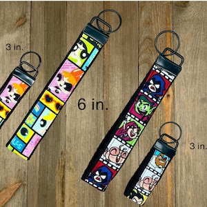 May include: Keychains featuring cartoon character designs. One keychain is 6 inches long, the other two are 3 inches long. The keychains have a black strap with a metal ring and a black top. The keychains are on a wooden surface.