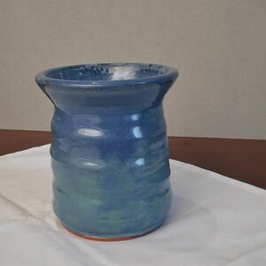 May include: A handmade ceramic vase with a blue and green glaze. The vase has a cylindrical shape with a slightly flared rim and a textured surface. The base is a reddish-orange color. The vase is sitting on a white cloth.