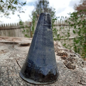 May include: A handmade, dark blue ceramic vase with a conical shape. The vase has a smooth, glossy finish with darker hues at the base and a lighter shade towards the top. It is displayed on a weathered, textured wooden surface.
