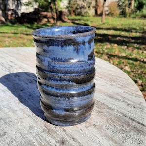 May include: A handmade ceramic tumbler with a blue and black glaze. The cylindrical cup features horizontal ridges and a glossy finish. The pottery is displayed on a weathered wooden surface, with a blurred outdoor background.