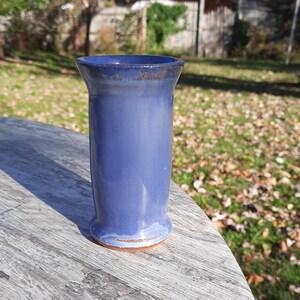 May include: A handmade, blue ceramic vase with a cylindrical shape and a slightly flared rim. The vase has a smooth, glossy finish and sits on a wooden surface. The vase is approximately 6 inches tall.
