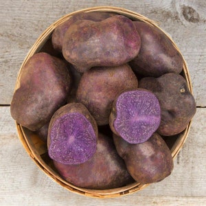 May include: A woven basket filled with purple potatoes, some whole and others cut in half to reveal their vibrant purple flesh. The potatoes have a dark purple skin and are arranged on a light wooden surface.