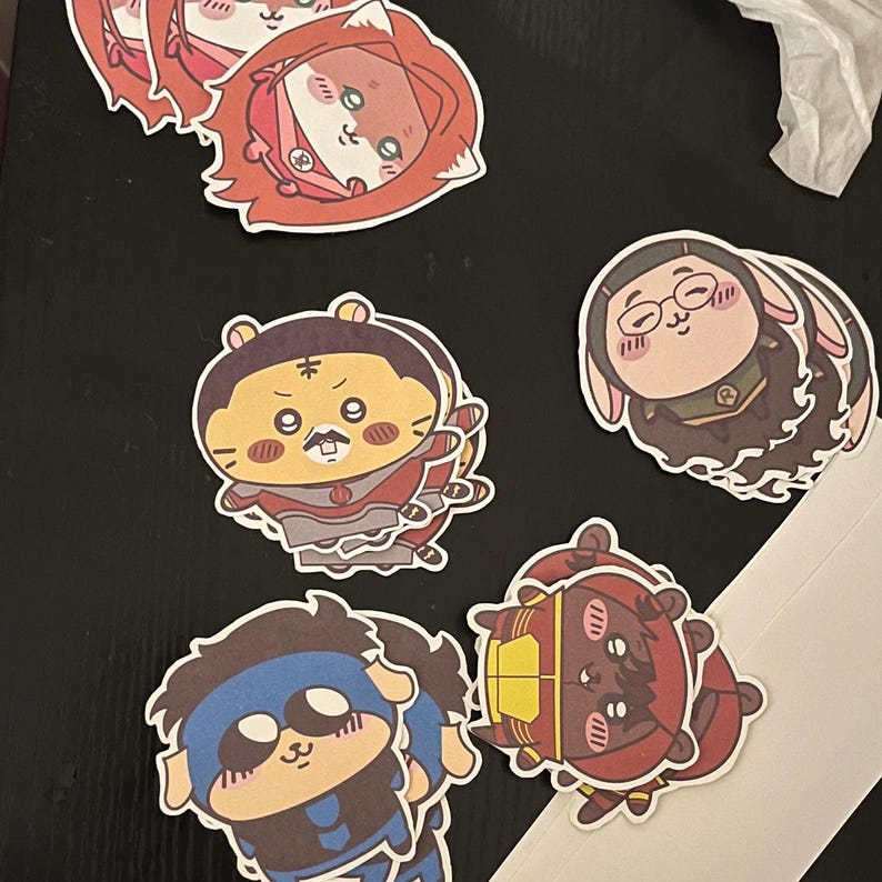 May include: A collection of colorful cartoon stickers featuring various animal characters. The stickers showcase a range of designs, including a fox, a tiger, a bunny, and other characters, each with unique outfits and expressions. The stickers are on a dark surface.