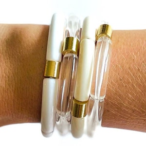 May include: A set of four white and clear bangle bracelets with gold accents. The bracelets are stacked on a person's wrist.