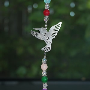 Könnte beinhalten: Ein silberner Kolibri-Ausschnitt-Anhänger hängt an einer Perlenkette mit einem Kristall-Tropfen am Ende. Die Perlen sind in Regenbogenfarben, darunter Grün, Rosa, Lila und Blau.