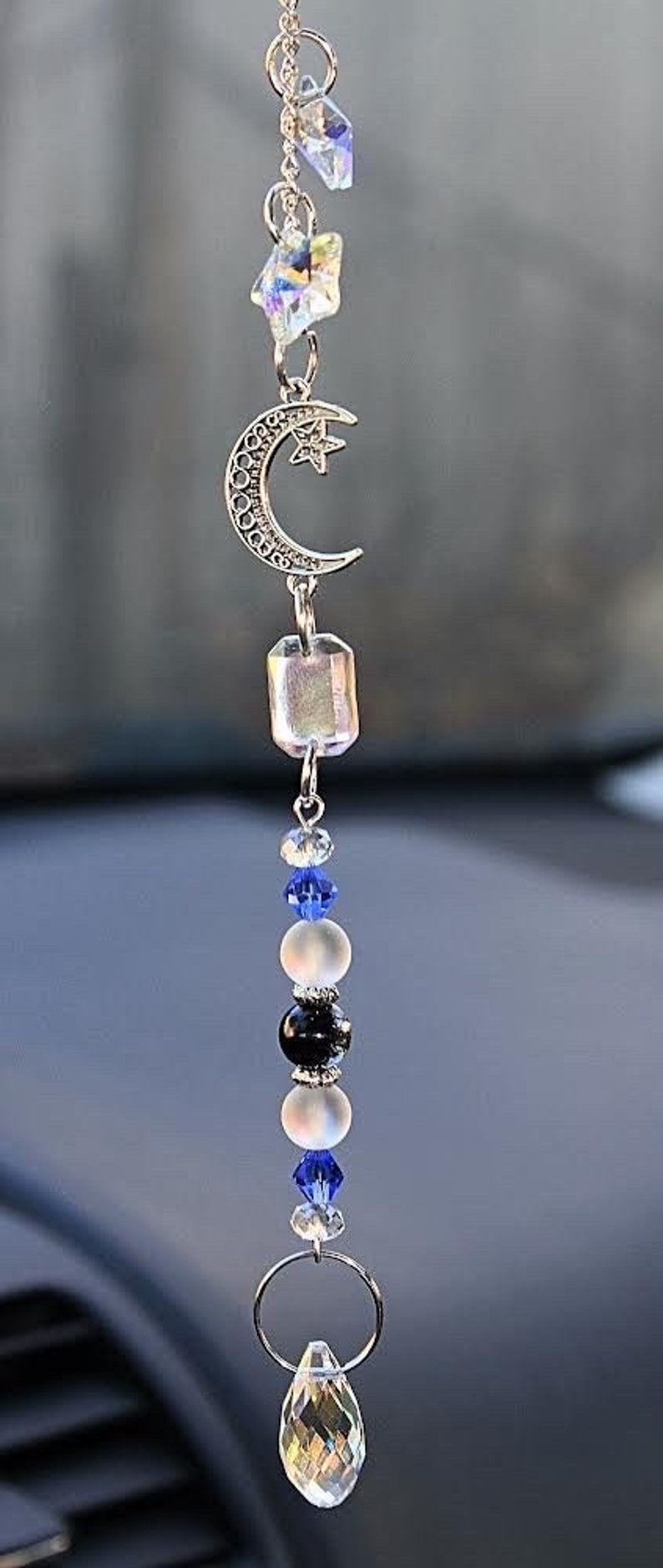 May include: A silver car charm featuring a crescent moon, star, and crystal beads. The charm hangs from a silver chain and has a clear crystal teardrop at the bottom.